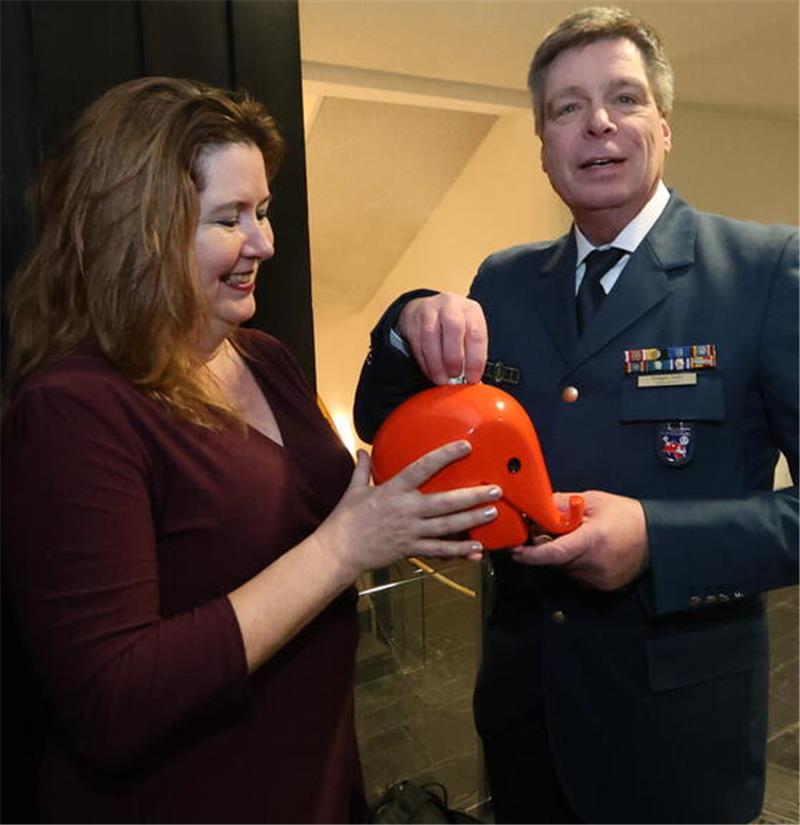 Eine Frau in dunkelrotem Kleid und ein Mann in Uniform mit Abzeichen halten gemeinsam eine orangefarbene Elefanten-Spardose aus Kunststoff.
