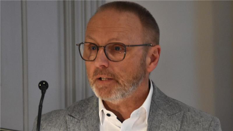 Mann mit Brille, weißem Hemd und grauem Sakko vor grauer Wand stehend