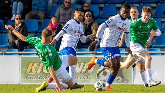 Fußballspiel mit zwei Spielern in weißen Trikots und zwei Spielern in grünen Trikots, einer grün gekleideten Person im Grätscheinsatz auf dem Spielfeld