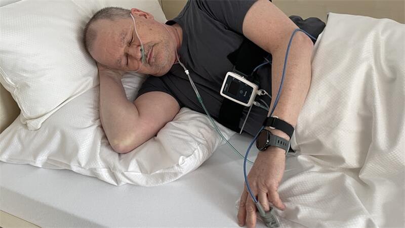 Ein Mann liegt im Bett. Für die Polygrafie werden die Patienten vor dem Schlafengehen mit einem Nasenschlauch, zwei Gurten und einem Fingersensor ausgestattet.
