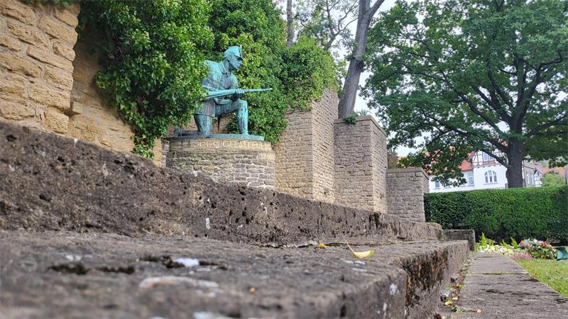 Statue eines Soldaten mit Gewehr auf einem gemauerten Sockel vor einer Steinmauer und Bäumen