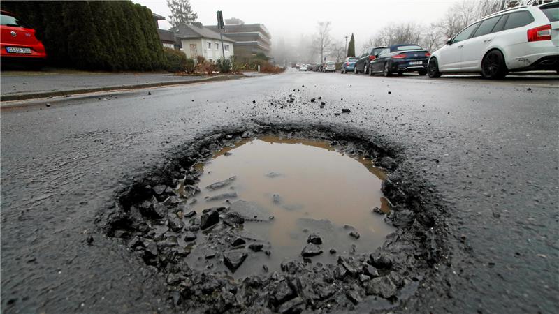 Großes Schlagloch mit Wasserfüllung auf einer Asphaltstraße in einem Wohngebiet mit geparkten Autos und Häusern im Hintergrund.
