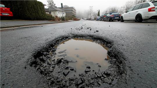 Großes Schlagloch mit Wasserfüllung auf einer Asphaltstraße in einem Wohngebiet mit geparkten Autos und Häusern im Hintergrund.