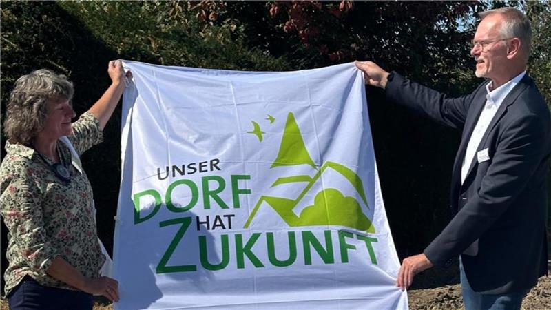 Nachbarschaftshilfe und Dorfgemeinschaft: Engagement im Nordharz Zwei Personen halten ein weißes Banner mit grünem Schriftzug und stilisierter Landschaft, auf dem steht 'Unser Dorf hat Zukunft'