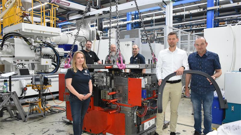 In einer Industriehalle stehen eine Frqau und fünf Männer an Maschinen, zwei halten ein Kunststoff-Autoteil in die Höhe.