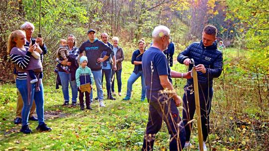 Eine Gruppe von Menschen im Wald, die zwei Personen beim Pflanzen eines Baumes zusehen.