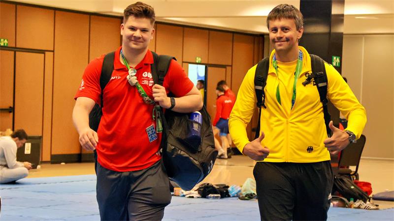 Zwei Männer mit Sporttaschen gehen nebeneinander durch einen Innenraum. Einer trägt ein rotes T-Shirt, der andere eine gelbe Jacke und zeigt beide Daumen hoch.