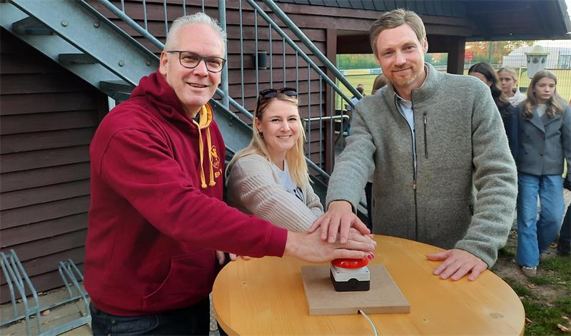 Drei Personen stehen an einem runden Holztisch und drücken gemeinsam einen großen roten Knopf, im Hintergrund weitere Personen und ein Holzgebäude mit Treppe