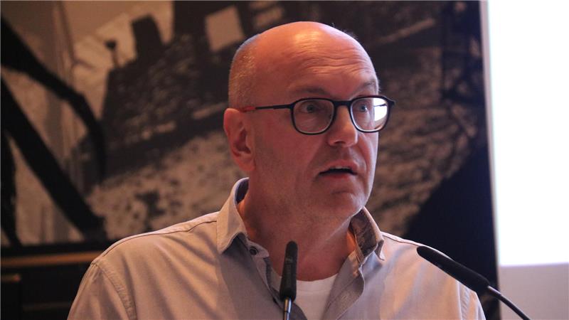 Wie die GZ den Aufstieg Adolf Hitlers feierte Mann mit Brille und hellblauem Hemd steht vor Mikrofonen, im Hintergrund unscharfe Wandgestaltung und Projektionsfläche.