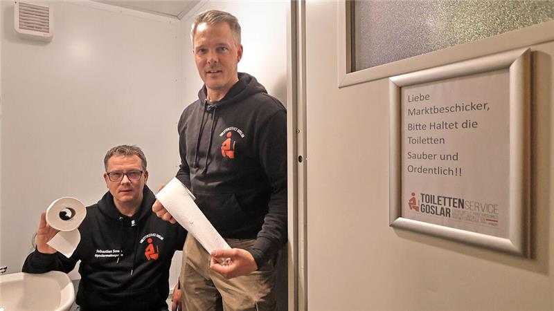 Zwei Männer in schwarzen Kapuzenpullovern in einem kleinen Raum, einer hält Toilettenpapier, an der Wand ein Schild mit der Aufschrift 'Liebe Marktbesicker, bitte haltet die Toiletten sauber und ordentlich!'