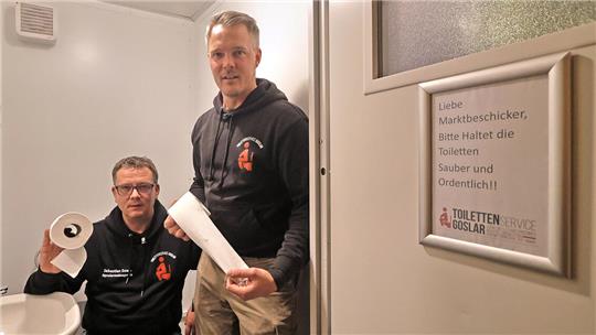 Zwei Männer in schwarzen Kapuzenpullovern in einem kleinen Raum, einer hält Toilettenpapier, an der Wand ein Schild mit der Aufschrift 'Liebe Marktbesicker, bitte haltet die Toiletten sauber und ordentlich!'