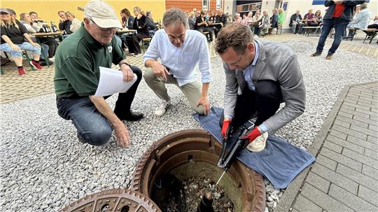 Drei Männer knien vor einem Loch im Boden, aus dem ein Stück Metall herausragt. Einer drückt Fett aus einer Speitze auf die Konstruktion. 