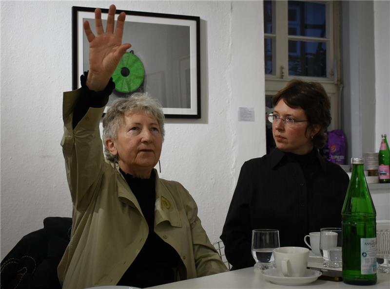 Zwei Personen sitzen an einem Tisch mit Gläsern, Tassen und einer grünen Flasche, eine Person hebt die Hand.