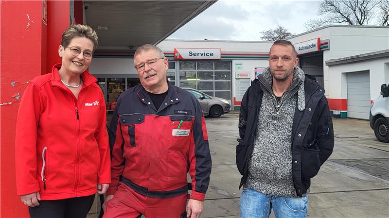Drei Personen stehen vor einer Werkstatt mit Service- und Autoschildern, zwei tragen rote Arbeitskleidung, eine Person trägt eine graue Strickjacke und blaue Jeans.