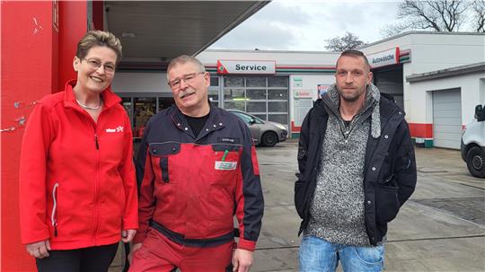 Drei Personen stehen vor einer Werkstatt mit Service- und Autoschildern, zwei tragen rote Arbeitskleidung, eine Person trägt eine graue Strickjacke und blaue Jeans.