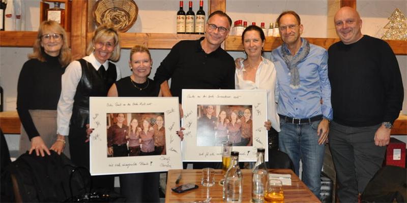 Gruppe von sieben Personen steht in einem Raum mit Weinflaschen und Dekoration, zwei halten gerahmte Fotos.