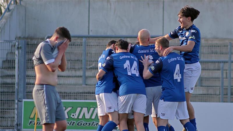 Fußballspieler in blauen Trikots feiern auf dem Spielfeld, während ein Spieler in grauem Trikot mit den Händen das Gesicht bedeckt steht
