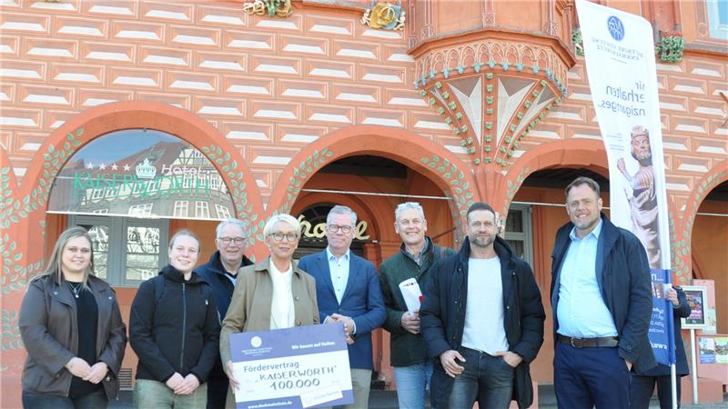 Gruppe von acht Personen vor einem Gebäude mit dekorativer Fassade und einem Banner mit Text
