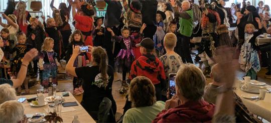Viele Menschen in einem Saal feiern und tanzen, einige Kinder tragen bunte Kostüme, im Hintergrund Banner mit den Worten 'Herzlich Willkommen! Harzklub e.V.' und 'Harz Energie'
