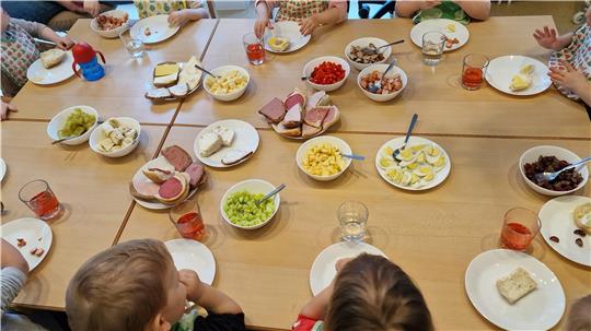 Kinder sitzen an einem Tisch mit Tellern und Schalen voller verschiedener Lebensmittel wie Brot, Wurst, Käse, Gemüse und Getränken.