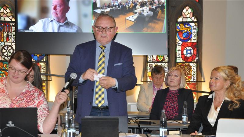 Mehrere Personen sitzen an einem Tisch in einem Raum mit bunten Glasfenstern, eine Person steht und hält ein Stück Papier, eine weitere Person hält ein Mikrofon