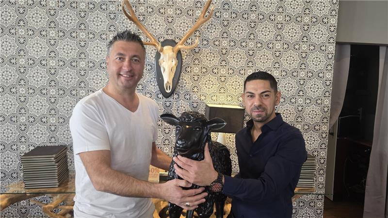 Zwei Männer mit kurzärmligem Hemd halten gemeinsam eine schwarze Ziegenfigur vor einer Wand mit gemustertem Tapetenhintergrund und einem an der Wand montierten Geweihschädel