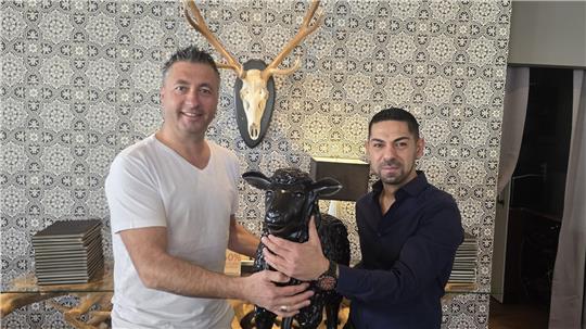 Zwei Männer mit kurzärmligem Hemd halten gemeinsam eine schwarze Ziegenfigur vor einer Wand mit gemustertem Tapetenhintergrund und einem an der Wand montierten Geweihschädel