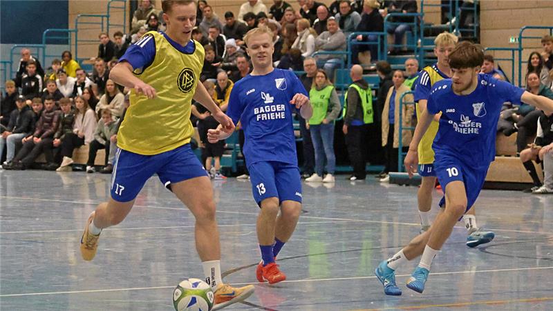 Mehrere Jugendliche spielen Hallenfußball, zwei tragen blaue Trikots, einer ein gelbes Leibchen, und ein Spieler führt den Ball.