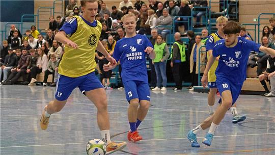 Mehrere Jugendliche spielen Hallenfußball, zwei tragen blaue Trikots, einer ein gelbes Leibchen, und ein Spieler führt den Ball.
