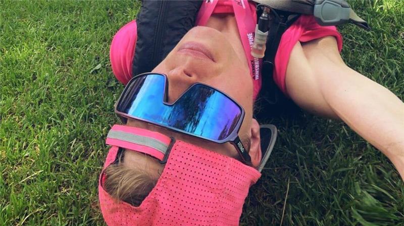 Person mit rosa Cap, schwarzer Weste und Sportbrille liegt auf dem Rücken auf einer Wiese mit ausgestreckten Armen.