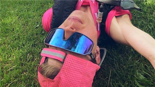 Person mit rosa Cap, schwarzer Weste und Sportbrille liegt auf dem Rücken auf einer Wiese mit ausgestreckten Armen.