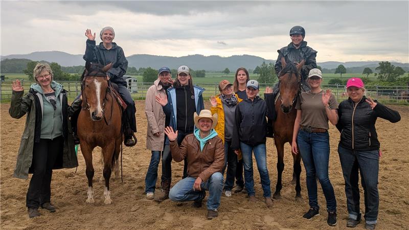 Gruppe von Menschen, zwei reiten auf Pferden, alle winken auf einem Reitplatz mit Bergen im Hintergrund