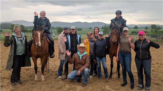 Gruppe von Menschen, zwei reiten auf Pferden, alle winken auf einem Reitplatz mit Bergen im Hintergrund