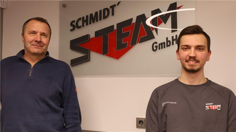Zwei Männer stehen vor einer Wand mit einem großen Logo, das den Schriftzug 'SCHMIDT STEAM GmbH' zeigt