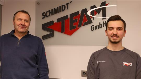 Zwei Männer stehen vor einer Wand mit einem großen Logo, das den Schriftzug 'SCHMIDT STEAM GmbH' zeigt