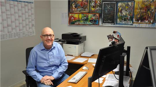 Person in blauem Hemd sitzt an einem Schreibtisch mit Computer, Drucker und mehreren Papieren, im Hintergrund bunte Gemälde an der Wand und ein Kalender.