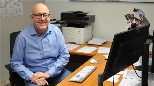 Person in blauem Hemd sitzt an einem Schreibtisch mit Computer, Drucker und mehreren Papieren, im Hintergrund bunte Gemälde an der Wand und ein Kalender.