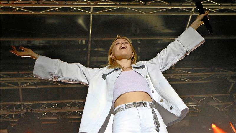 Frau mit ausgestreckten Armen auf einer Bühne vor einem Gerüst aus Metallträgern