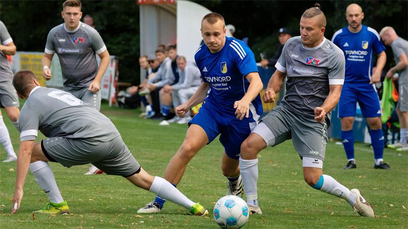 Fußballspieler in blauen und grauen Trikots kämpfen um den Ball auf einem Spielfeld mit Zuschauern im Hintergrund