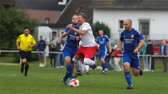 Zwei Fußballspieler in blauen Trikots und ein Spieler in weißem Trikot mit roter Hose kämpfen um den Ball auf dem Spielfeld, im Hintergrund Schiedsrichter und Zuschauer vor Wohnhäusern