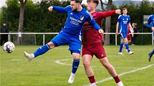 Zwei Fußballspieler kämpfen um den Ball, einer in blauem Trikot hebt ein Bein zum Schuss, der andere in rotem Trikot versucht zu blocken
