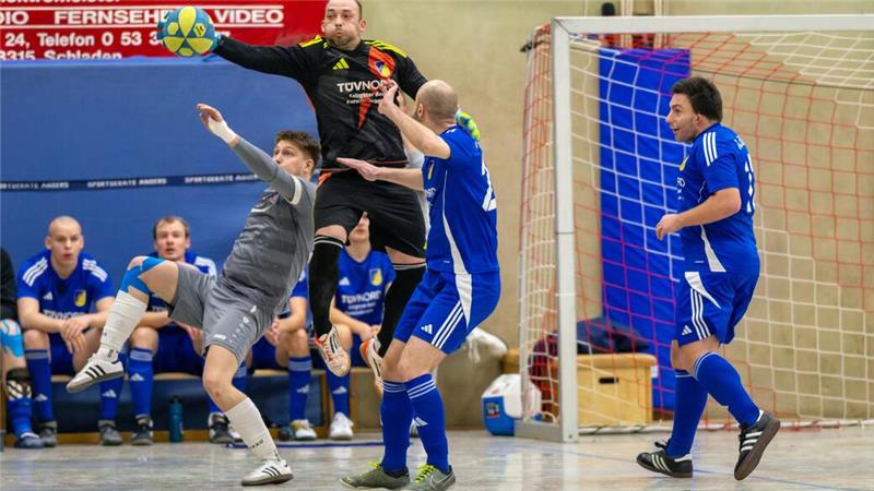 Mehrere Hallenfußballspieler in Aktion vor dem Tor, ein Spieler springt mit Ball in der Hand, andere verteidigen oder beobachten das Spiel.