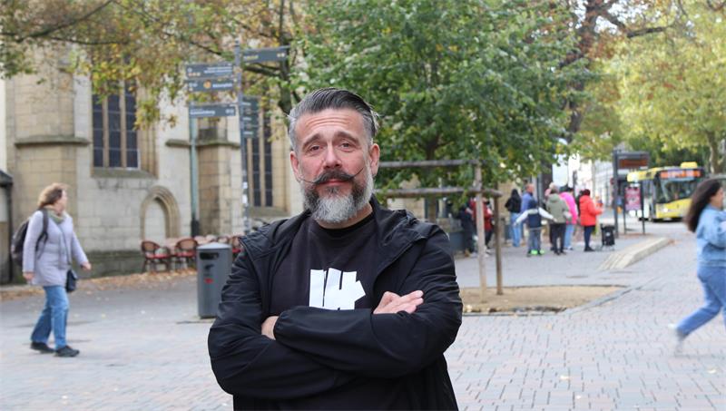 Mann mit grauem Bart und schwarzer Jacke steht mit verschränkten Armen auf einer belebten Straße mit Bäumen und Gebäuden im Hintergrund.