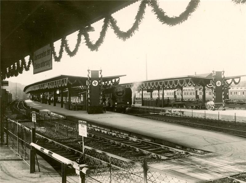 Schwarz-weiße Aufnahme eines mit Hakenkreuzfahnen und Girlanden geschmückten Bahnhofs mit mehreren Bahnsteigen und Gleisen.