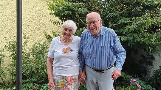 Älteres Paar steht lächelnd in einem Garten, der Mann stützt sich auf einen Gehstock, im Vordergrund hält eine Person ein Smartphone zum Fotografieren