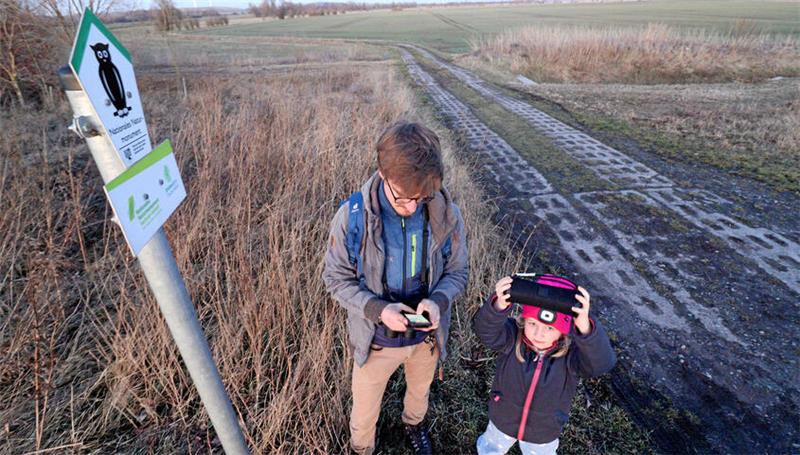 Zwei Personen stehen an einem Weg aus Betonplatten, neben ihnen ein Schild, das aufs Grüne Band hinweist.