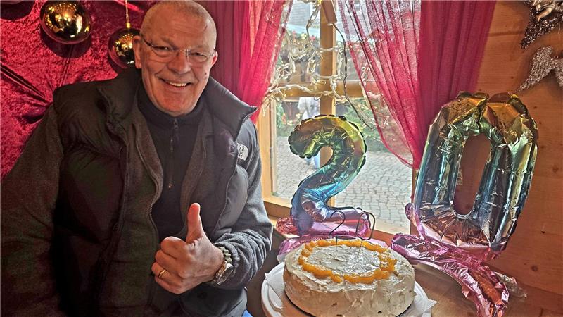 Mann mit Daumen hoch neben rundem Kuchen mit orangem Zuckerdekor und gold-grünen Luftballons in Form der Zahl 20 vor Fenster mit roten Vorhängen.