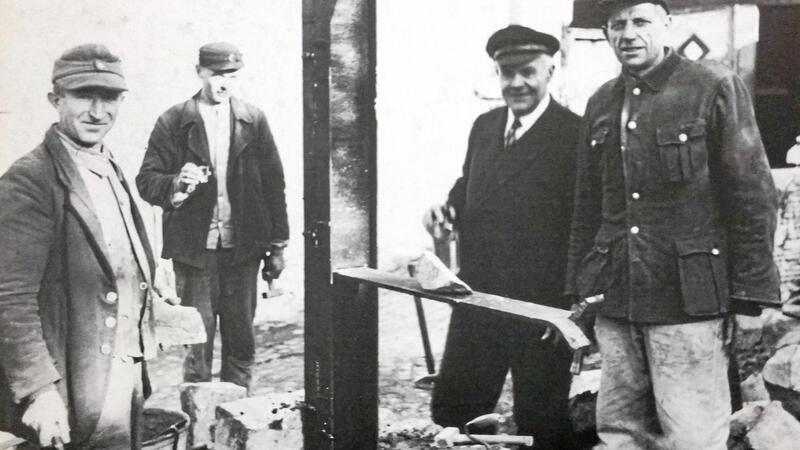 Vier Männer in Arbeitskleidung stehen um eine Baustelle mit Ziegelsteinen und einem Brett auf einem Pfosten