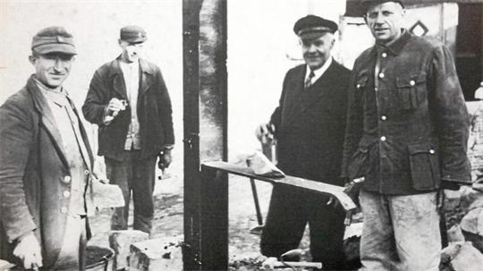 Vier Männer in Arbeitskleidung stehen um eine Baustelle mit Ziegelsteinen und einem Brett auf einem Pfosten
