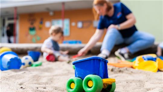 Spielzeugauto mit grünen Rädern im Sandkasten und unscharf spielendes Kind mit Erwachsenem im Hintergrund